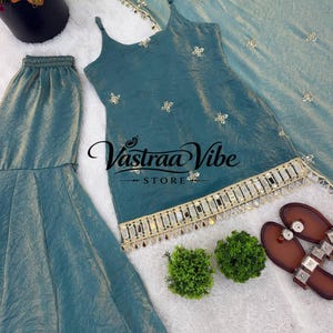 May include: A teal sleeveless tunic with thin straps, featuring gold floral embroidery and a decorative hem with mirrors and hanging beads. The set includes matching flared trousers and a coordinating dupatta, with the text Vastraa Vibe STORE.