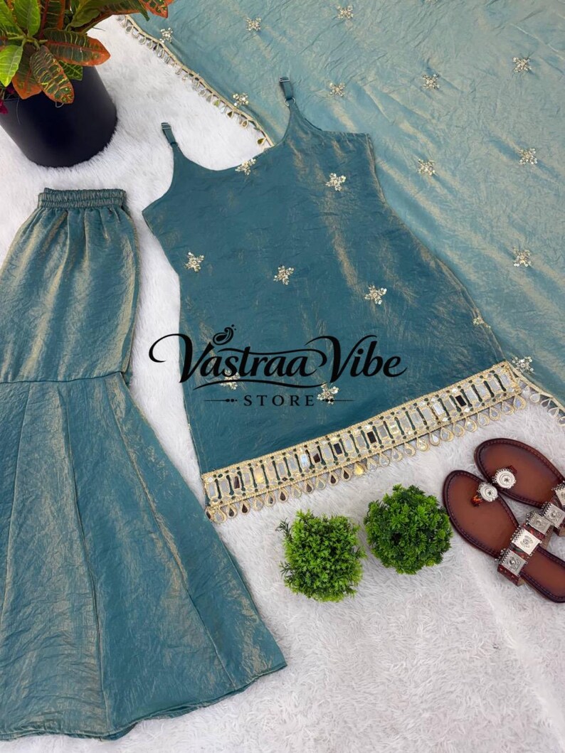 May include: A teal sleeveless tunic with thin straps, featuring gold floral embroidery and a decorative hem with mirrors and hanging beads. The set includes matching flared trousers and a coordinating dupatta, with the text Vastraa Vibe STORE.