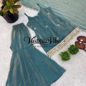 May include: A teal sleeveless top with thin straps, matching flared pants, and a coordinating dupatta, all featuring small gold floral embroidery. The top hem is decorated with a gold fringe trim. The text Vastraa Vibe STORE is visible.