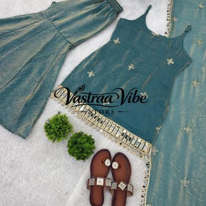 May include: A teal sleeveless top with thin straps, matching wide-leg pants, and a long scarf, all featuring gold embroidery and mirror accents. The top displays the text Vastraa Vibe Store. A pair of brown flat sandals with metallic embellishments.