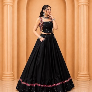 May include: A black two-piece set consisting of a sleeveless crop top and a floor-length skirt. The top features thin straps with colorful embroidery and small mirrors. The skirt has a wide hem with matching embroidery, mirrors, and side pockets.
