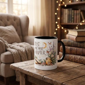 Op de afbeelding: Een witte keramische mok met een zwarte handgreep en rand, voorzien van de tekst "JUST ONE MORE CHAPTER" in gouden letters, vergezeld van illustraties van een maansikkel, vlinders, een kaars, een boek en een boeket lavendel en wilde bloemen.