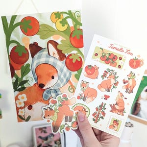 May include: A rectangular art print and a sticker sheet featuring illustrations of orange foxes with tomatoes. The print shows a fox wearing a blue gingham headscarf among tomato vines. The sticker sheet includes the text Tomato Fox and various fox designs.