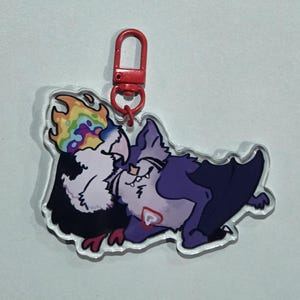 May include: A clear acrylic charm featuring two cartoon characters. One is a black and white bird with a rainbow flame on its head, and the other is a purple bat-like creature. The charm has a red clasp.