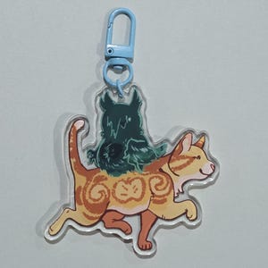 May include: A clear acrylic keychain featuring a cartoon cat with orange and brown stripes, carrying a dark green creature on its back. The keychain has a light blue clasp.