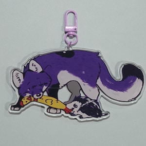 May include: A purple and white fox-like character keychain with a lobster clasp. The fox is holding a yellow object with a red design. A black and white character is on the bottom. The keychain is attached to a purple clasp.