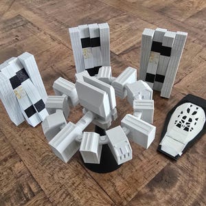 May include: A collection of white and black 3D-printed objects, including a central articulated structure, three rectangular panels, and a black and white knee pad. The objects are arranged on a wooden surface.