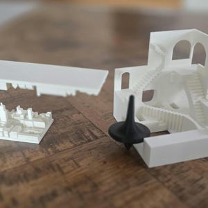 Puede incluir: Maqueta arquitectónica blanca con una sección recortada que revela un paisaje urbano. Un trompo negro se encuentra cerca de una estructura blanca con múltiples escaleras. La maqueta está sobre una superficie de madera.