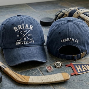 May include: Two navy blue baseball caps with white text and a hockey theme. One cap reads "Briar Hawks University" with crossed hockey sticks. The other cap has "Graham 44" on the back. Hockey stick, puck, and pins are also visible.