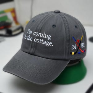May include: A gray baseball cap with the embroidered text "I'm coming to the cottage." The cap features a curved brim and a small embroidered design of crossed hockey sticks with the numbers "24" and "81".