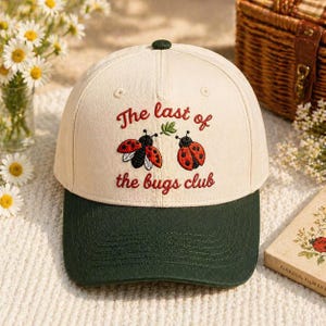 Puede incluir: Gorra de béisbol color crema con visera verde oscuro. La gorra presenta mariquitas rojas bordadas y el texto "The last of the bugs club" en escritura roja. Un pequeño botón verde está en la parte superior.