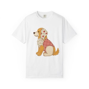 Puede incluir: Una camiseta blanca de manga corta con un gráfico central de un perro sentado. El perro está compuesto por varios parches de tela, incluyendo un patrón de cuadros vichy rojo y blanco, un estampado de fresas, flores y puntos color crema.