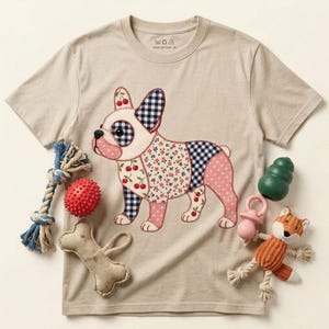 Puede incluir: Una camiseta beige de manga corta con un gráfico de bulldog francés estilo patchwork con patrones de cerezas, cuadros vichy y flores. La camiseta está rodeada de varios juguetes para mascotas, incluyendo una cuerda, una pelota roja y un zorro.