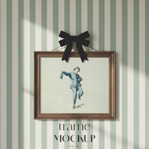 May include: A framed artwork depicting a figure in a blue costume, suspended against a striped wall. A black bow is tied above the frame. The frame is brown. The text "frame MOCKUP" is at the bottom.