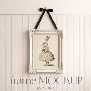 May include: A framed print featuring a vintage illustration of a rabbit in a floral dress. The white ornate frame is suspended by a black ribbon and bow. The text "frame MOCKUP" is displayed at the bottom of the image.