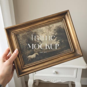 Puede incluir: Un marco rectangular dorado con un borde detallado, que contiene una imagen de paisaje con un caballo blanco. El marco tiene las palabras "frame MOCKUP" en blanco. La imagen la sostiene una persona.