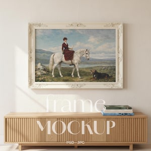 May include: A framed painting of a woman on a white horse with a dog in a landscape. The painting is in a white ornate frame. The frame is hanging above a light wood cabinet. The word "frame" and "MOCKUP" are displayed on the image.