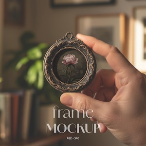 Puede incluir: Un pequeño marco redondo y ornamentado, negro con detalles dorados, que contiene una pintura de una peonía rosa. El marco está sostenido en una mano. El texto "frame MOCKUP" está en la parte inferior de la imagen.