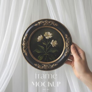 May include: Round black framed artwork featuring a floral painting with white flowers and green leaves. The frame has gold-colored ornate detailing. The words "frame MOCKUP" are printed in white at the bottom of the image.