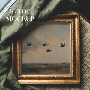 Op de afbeelding: Een ingelijst vintage landschapsschilderij met vier vliegende vogels tegen een bewolkte lucht. De goudkleurige lijst heeft sierlijke details. Een groene stof bedekt het schilderij gedeeltelijk. De tekst "frame MOCKUP" is zichtbaar.