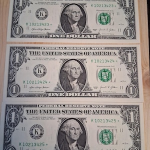 May include: Three United States one-dollar bills. Each bill features a portrait of George Washington and the text "ONE DOLLAR". The bills are green and black with intricate designs and serial numbers. The bills are on a light brown wooden surface.