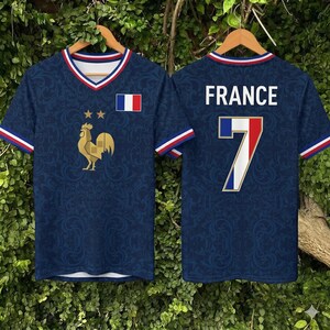 May include: Two navy blue soccer jerseys on wooden hangers. The front jersey features a gold rooster emblem, a French flag patch, and the number 7 on the back. Both jerseys have a V-neck collar with red, white, and blue trim.
