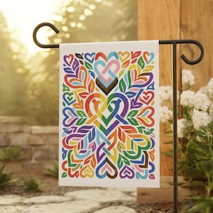 May include: A white garden flag featuring a colorful heart design with rainbow and Pride flag elements. The flag is held by a black metal stand, with a blurred background of greenery and a stone wall.