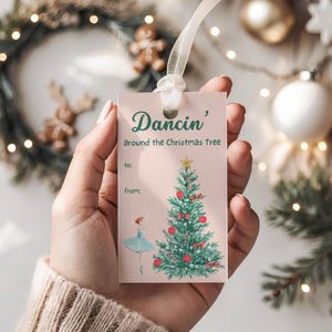 Könnte beinhalten: Ein rosa Geschenkanhänger mit dem Text "Dancin' around the Christmas Tree". Der Anhänger zeigt eine Aquarellillustration eines Weihnachtsbaums mit roten Kugeln und einer Ballerina. Ein weißes Band ist befestigt.