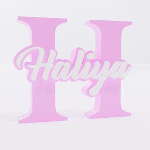 Puede incluir: Una letra "H" rosa tridimensional con el nombre "Haliya" en escritura blanca en el centro. Las letras son de color rosa pastel sólido, y el nombre está en una fuente cursiva.