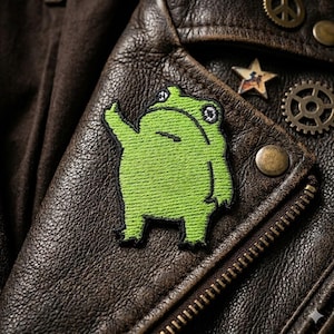 May include: A green embroidered frog patch on a brown leather jacket. The frog is facing left with its arm raised. The jacket has a zipper and metal embellishments.
