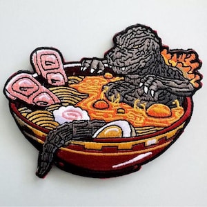 May include: Embroidered patch featuring Godzilla in a bowl of ramen. The patch has a red and yellow bowl filled with noodles, broth, and toppings. Godzilla is gray with white claws and is emerging from the ramen.