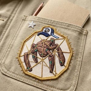 May include: Close-up of a tan pocket with a detailed embroidered patch featuring a brown insect-like creature wearing a blue cap. A small star pin and a notebook with a pencil are also visible.