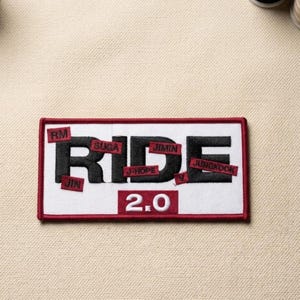 May include: A rectangular patch with a white background and a red border. The word "RIDE" is in large black letters, with smaller red rectangles above and below the letters, each with a name. The number "2.0" is below the word "RIDE".