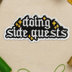 May include: Embroidered patch with the words "going side quests" in white script on a black background. The patch has a white border and yellow star accents. The patch is on a beige fabric surface.