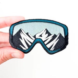 May include: Embroidered patch featuring a pair of ski goggles with a mountain range scene. The goggles have a teal frame and a black base, with white and black mountain peaks visible through the lenses. Ideal for jackets, bags, or other fabric items.