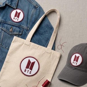 May include: A denim jacket, canvas tote bag, and gray baseball cap, each featuring a circular patch with the letters "BTS" and a stylized logo in burgundy and white. The items are arranged on a neutral surface.