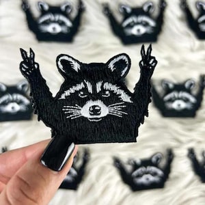 May include: Black embroidered raccoon patch with white accents, giving a peace sign with its paws. The patch is held up in front of a white, fluffy background, with several other raccoon patches visible.