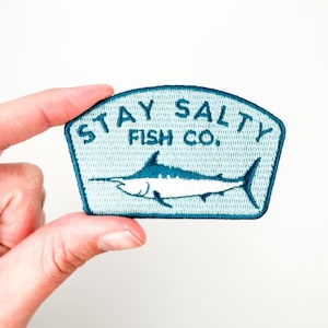 May include: A light blue embroidered patch with the text "STAY SALTY FISH CO." and a marlin fish design. The patch is held by a hand against a white background. The patch is a shield shape.