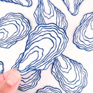 May include: White fabric with multiple blue embroidered oyster shell designs. The shells have a detailed, layered appearance, with each shell featuring a unique pattern of concentric lines. The design is set against a white background.