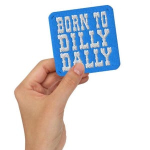 May include: A square, blue embroidered patch with the words "BORN TO DILLY DALLY" in white, block letters. The patch is held by a hand against a white background. The patch has a stitched border.