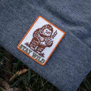 May include: Gray beanie with a patch featuring a brown Bigfoot holding a mug with steam rising. The patch has an orange border and the words "STAY WILD" in green. The beanie is made of a knitted material.