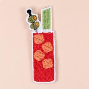 May include: Embroidered patch featuring a red cocktail with orange ice cubes, green celery, and olives on a toothpick. The patch has a white border and is designed to be ironed or sewn onto fabric. Perfect for adding a unique touch to clothing or accessories.