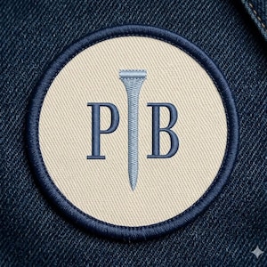 May include: Circular embroidered patch with a navy blue border. The patch features a light beige center with the letters "P" and "B" in navy blue on either side of a golf tee. The patch is set against a dark blue denim background.