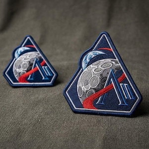 May include: Two embroidered patches with a space theme. Each patch features a gray moon with craters, a red curved line, and a blue and white planet. The patches are in a dark blue triangular shape with white and blue accents.