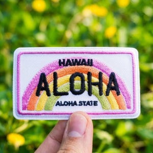 May include: Embroidered patch with the word "ALOHA" in black letters over a rainbow design. The patch reads "HAWAII" above the rainbow and "ALOHA STATE" below. The patch has a white background and a pink border.
