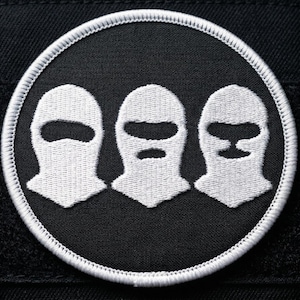 May include: Embroidered patch featuring three white ski masks on a black circular background, bordered by a white stitched edge. The masks have different eye and mouth openings. The patch is set against a black fabric.