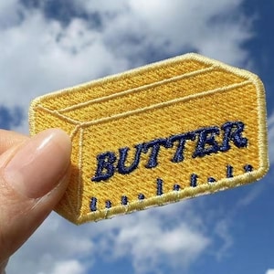 May include: Embroidered yellow patch in the shape of a butter stick. The word "BUTTER" is stitched in navy blue across the front. The patch is held against a blue sky background.