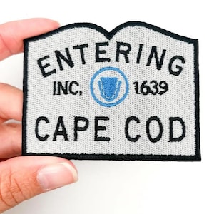 May include: Embroidered patch with the words "ENTERING CAPE COD" in black, along with "INC. 1639" and a blue emblem. The patch has a light gray background and a black border.