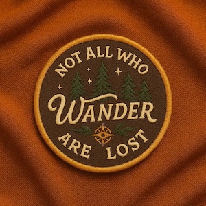 May include: Embroidered patch with the text "NOT ALL WHO WANDER ARE LOST" in cream-colored script and block letters. The patch features a brown background with green trees, stars, and a compass rose design, all framed by a gold border.