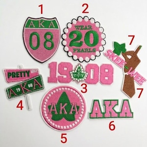 May include: A collection of embroidered patches in pink, green, and white. The patches feature text and symbols, including "AKA", "Wear 20 Pearls", and "Pretty Way". Various shapes include a shield, circle, and sign.